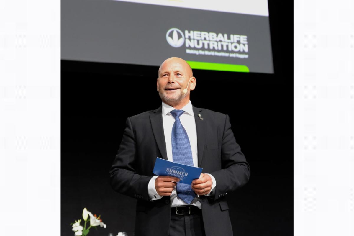 A person standing on a stage, holding a blue card. "herbalife nutrition" logo behind them.