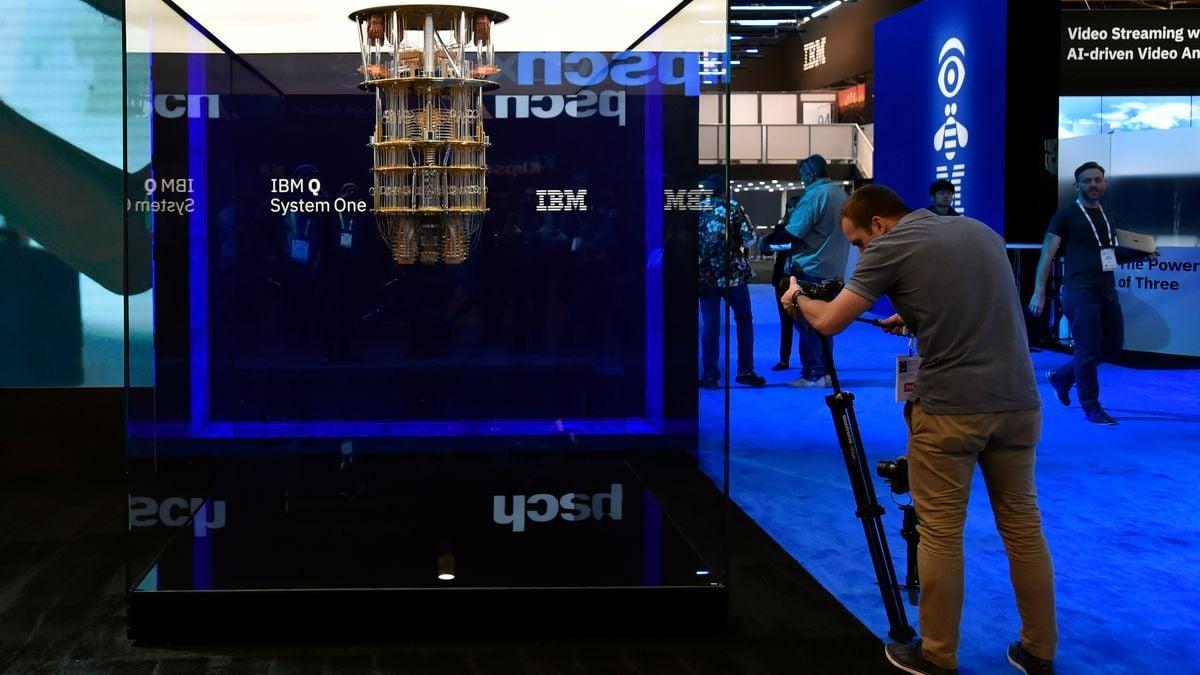 A person using a camera on a tripod to photograph a large device hanging from the ceiling of an glass enclosed space. "IBM SystemQ" on the back wall.