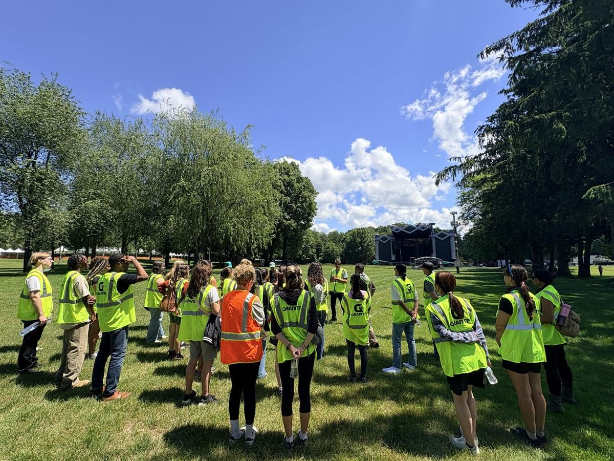 College program participants wearing high-visibility safety vests stand in a grassy field while receiving a festival operations briefing with an outdoor stage visible in the distance.