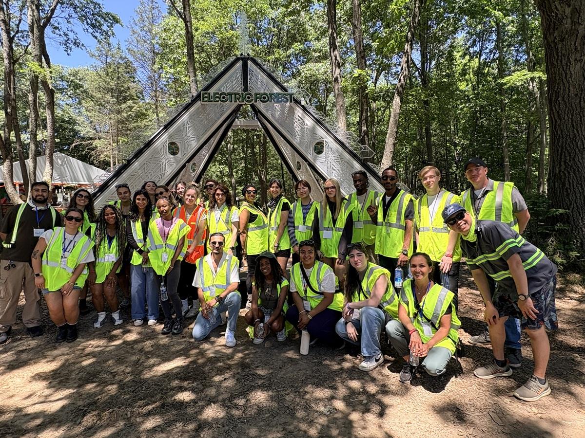 A group of college program participants wearing high-visibility safety vests poses for a photo beneath the Electric Forest entrance structure in a wooded festival setting surrounded by trees.