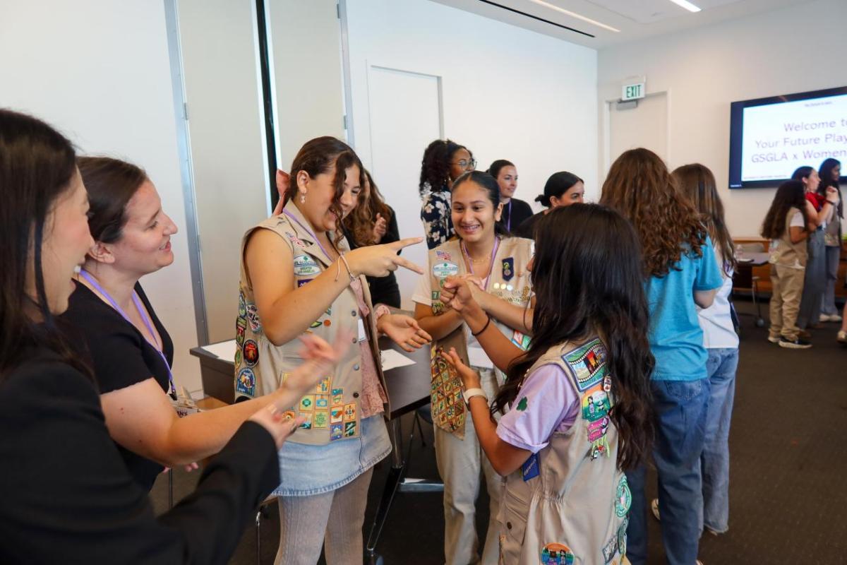 Participants stand in a circle indoors, engaging in an interactive group activity during a workshop, with a presentation screen visible in the background.