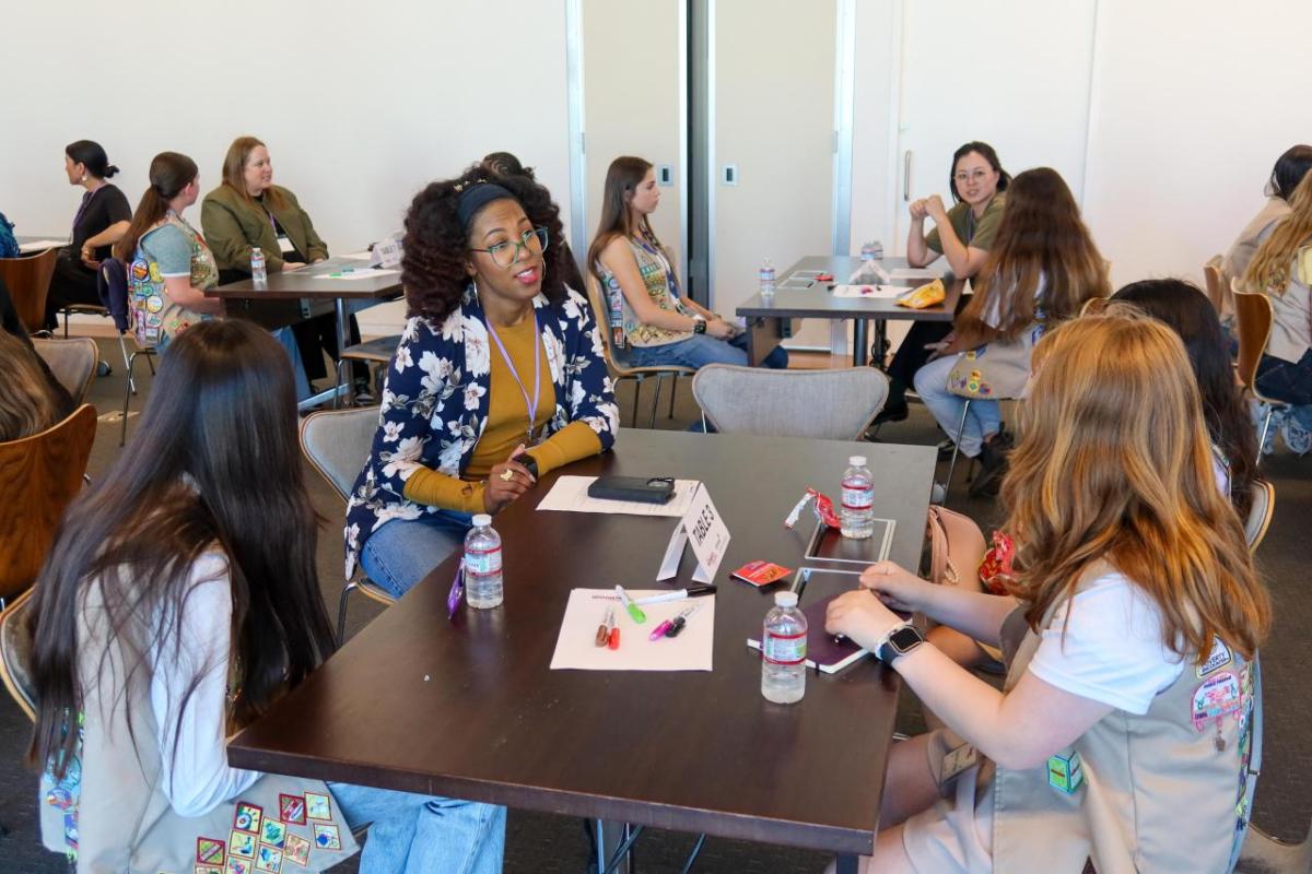 Participants sit around a table during a collaborative workshop, with notebooks, markers, and water bottles spread across the table in a modern meeting room.
