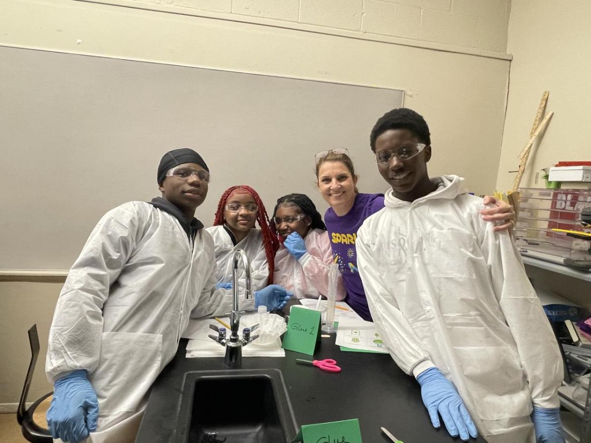 A woman in a purple Curiosity Labs™ MilliporeSigma t-shirt stands with students in lab equipment as they conduct experiments. 