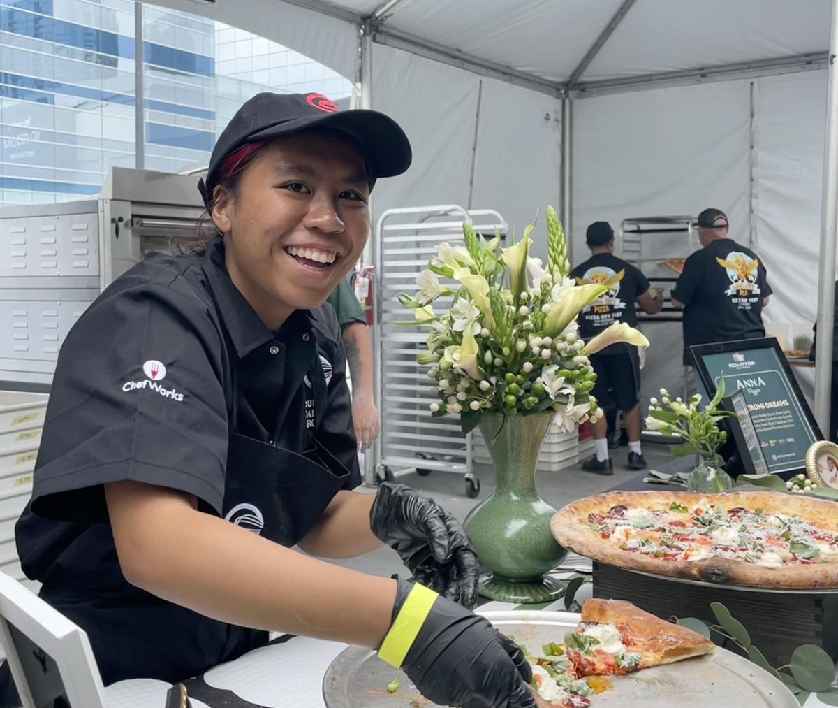 Food service worker wearing gloves prepares pizza slices at a festival booth, with a decorated pizza display on the counter.