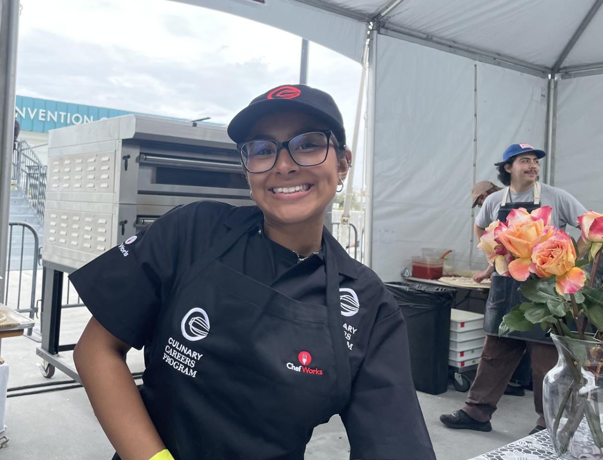 Culinary Careers Program participant stands at a pizza booth wearing a branded apron, with pizza ovens visible in the background.