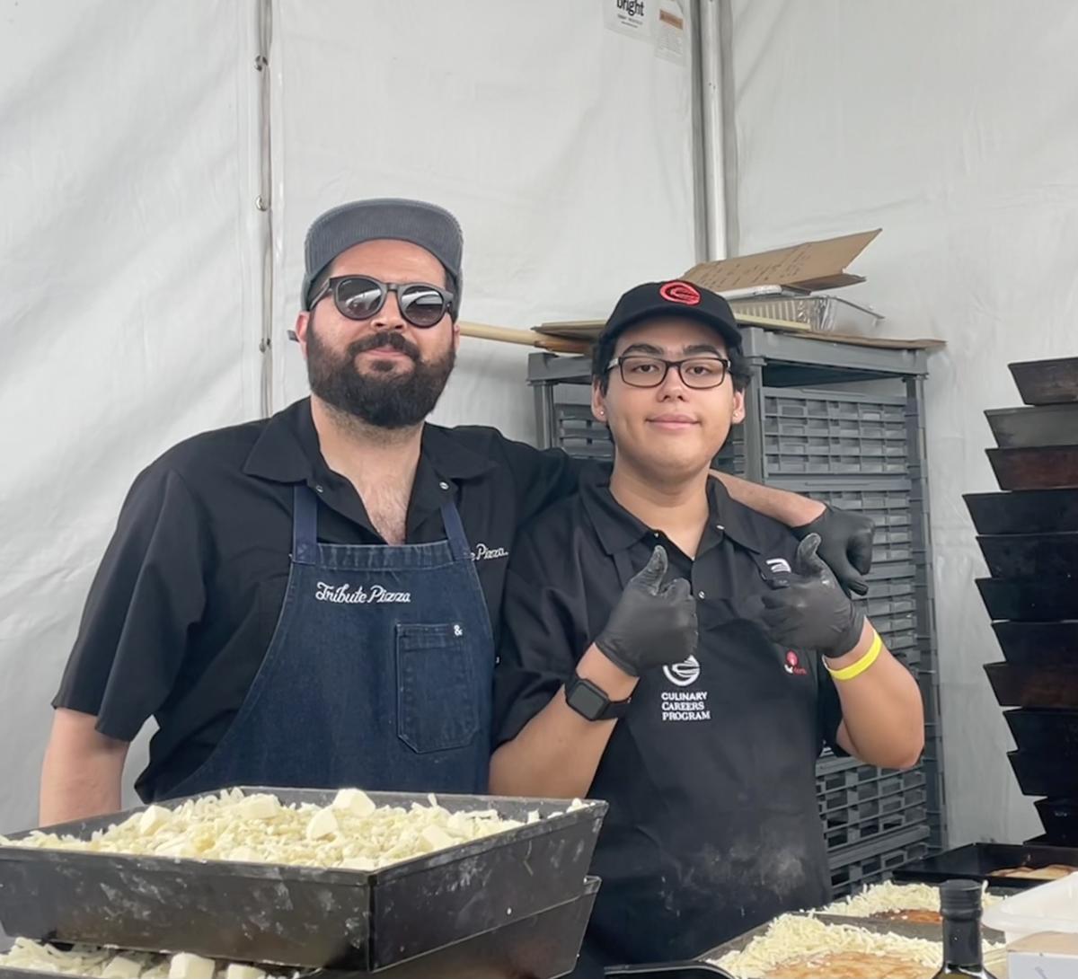 Two food service workers stand behind a pizza prep station with trays of shredded cheese inside a tent.