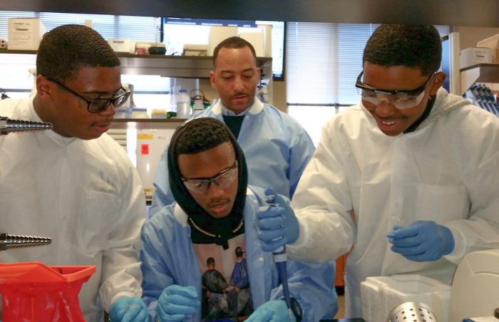 Dr. Arik King in a lab working with three young men of color. All are wearing lab coats and protective eye ware. 