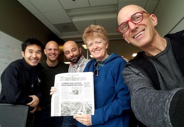 Shannon Whitmore with three men, holding a newspaper with a headline "Illumina Buys Startup"