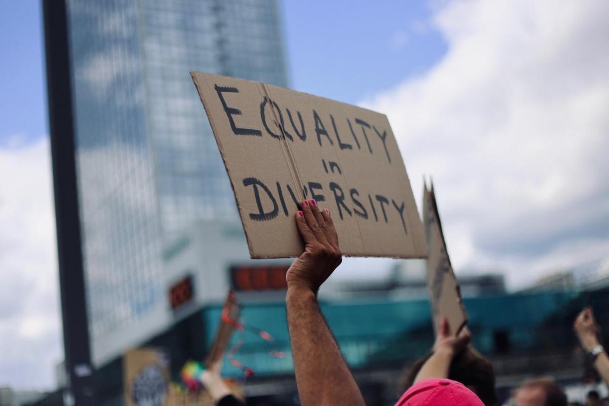 A hand holding up a cardboard sign "Equality in Diversity". A tall building in the distance.