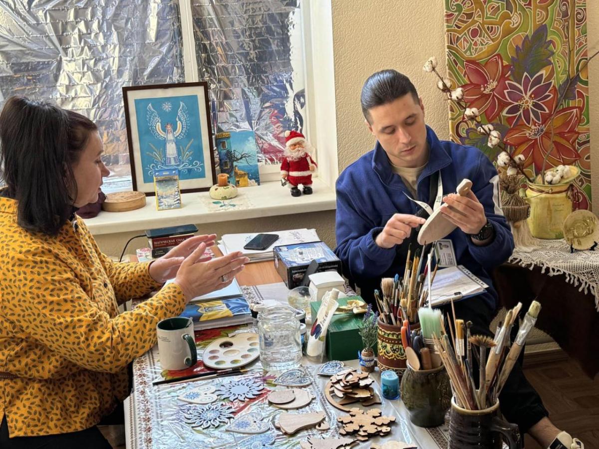 A woman in a yellow shirt gives a lesson to a man in a blue jacket. The are surrounded by painting materials.
