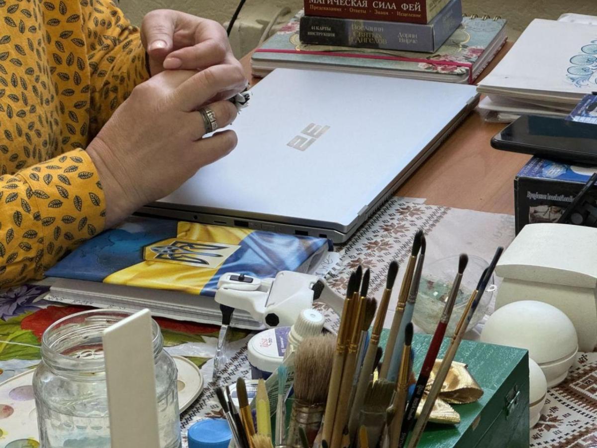 A woman's interlaced fingers hover over a closed laptop. Paintbrushes are on the desk.