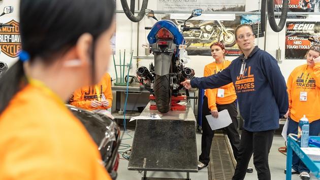 An adult pointing to a spot on a raised motorcycle as students look on.