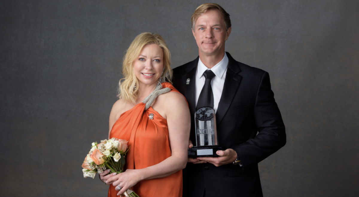 Two people stood next to each other smiling, one person holding flowers and the other holding an award