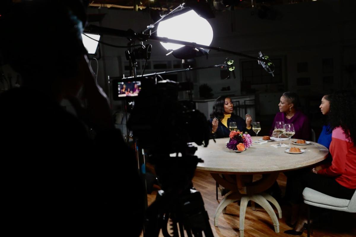 QVC Hosts Vanessa Herring, Jayne Brown, Monifa Days and Stacey Rusch are seated at a table with a television camera filming them while on-set at QVC Studios.