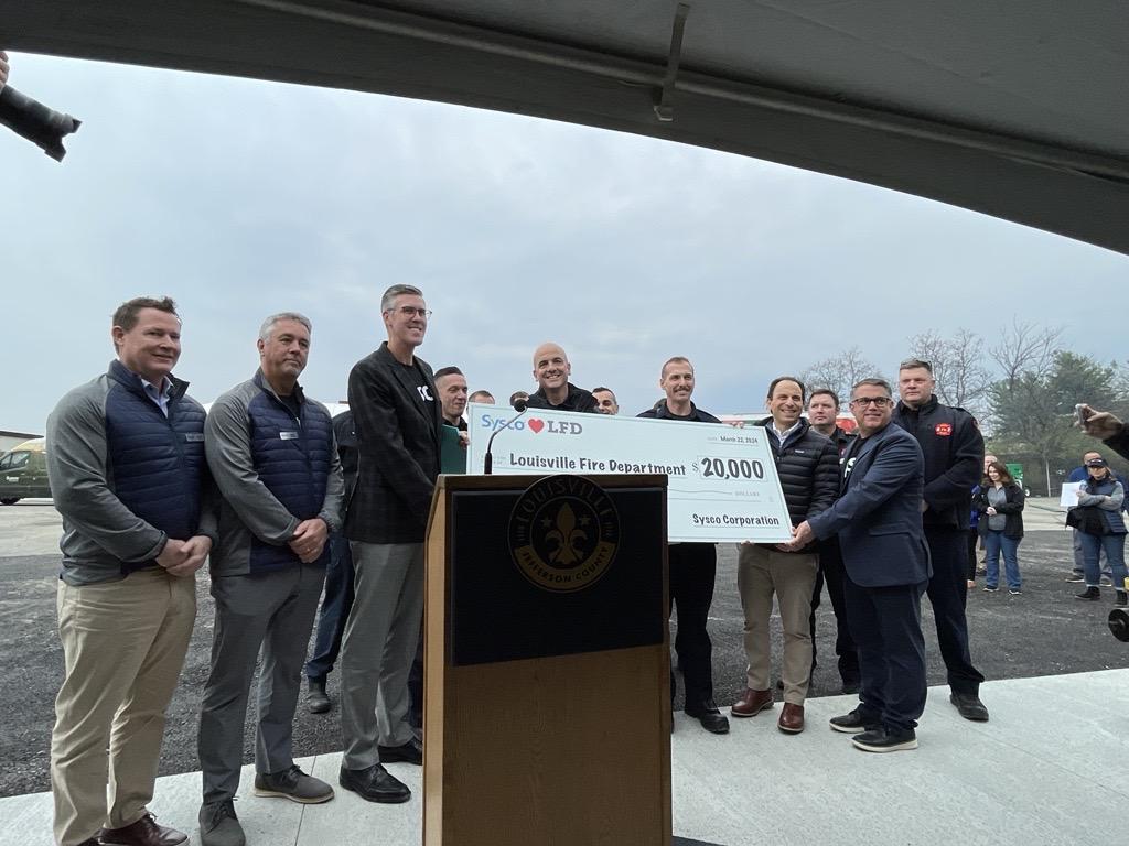 A group of people posed with a large check behind a podium outside.