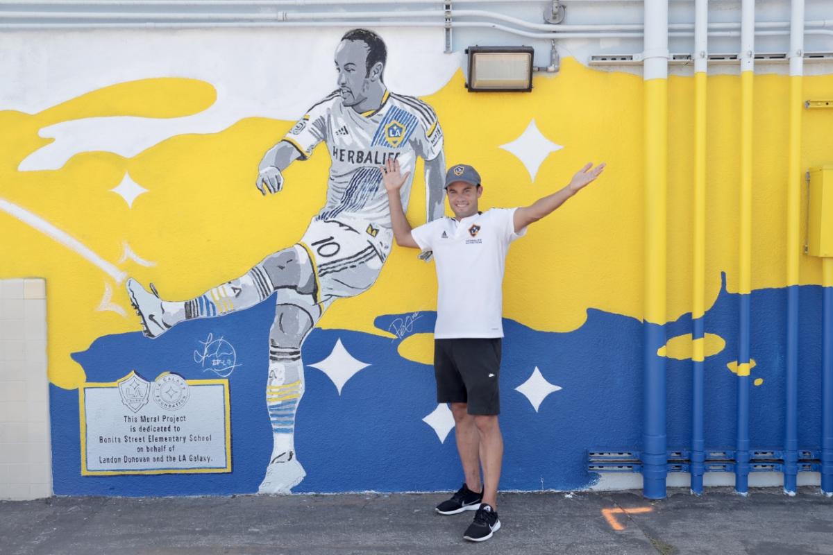 LA Galaxy player Landon Donovan poses with his arms outreached in front of a yellow, blue and white mural of him in a LA Galaxy jersey kicking a soccer ball. 