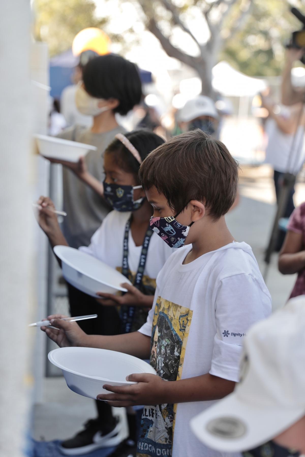 Elementary students wearing masks, each hold bowls of paint and brushes as they stand in a line and finish the LA Galaxy mural at their school. 