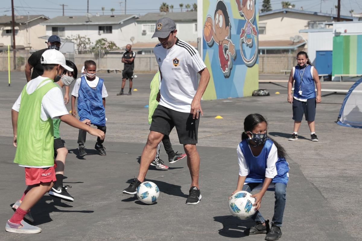 Students in green and blue penny jerseys kick a soccer ball around with LA Galaxy legend Landon Donovan on the blacktop of a local elementary school. 