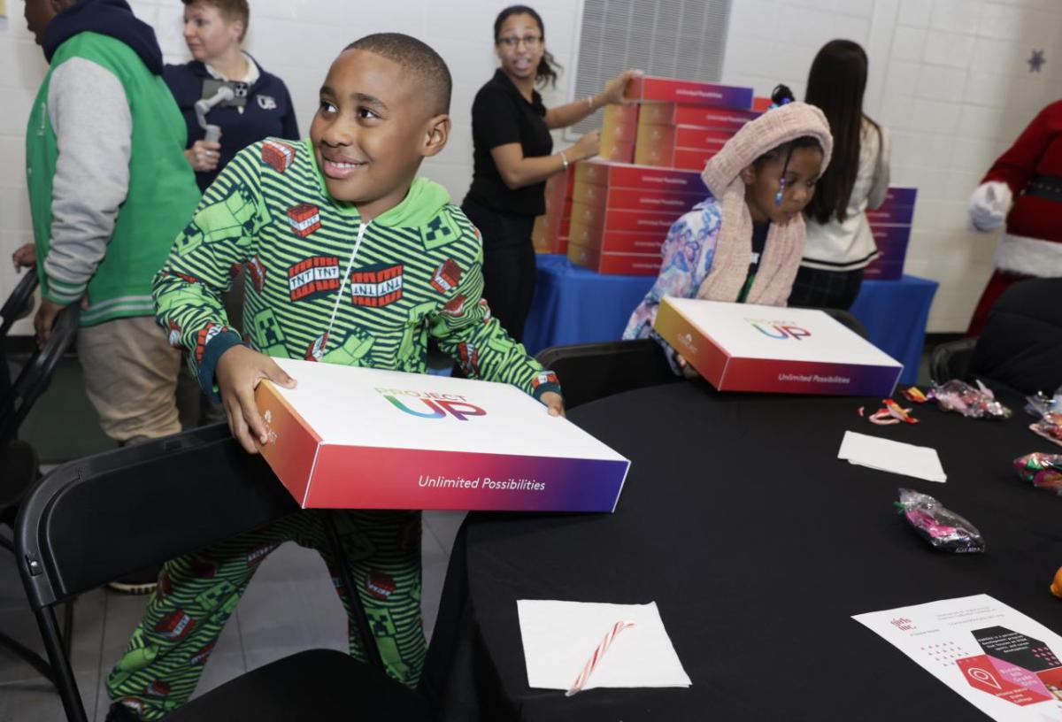 Excited children with boxes labeled "project UP". A stack of boxes behind them and a round black table in front .