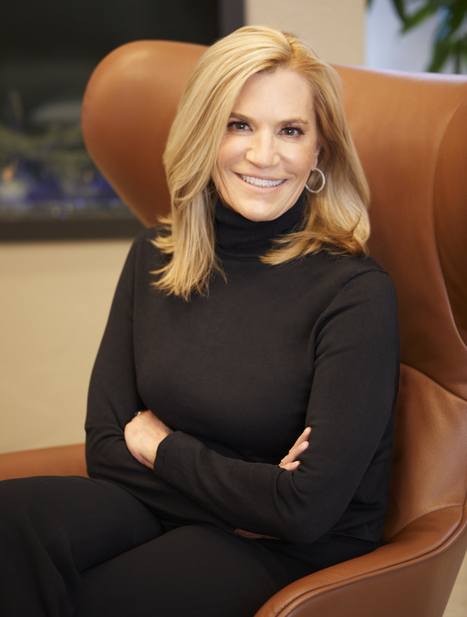 Laura Kohler sitting in a wingback leather chair, smiling, arms crossed, wearing a black turtleneck and pants.