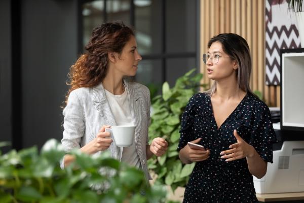 Two people talking, one with a white cup in their hand, the other has a phone. In an office setting with house plants in front and behind.