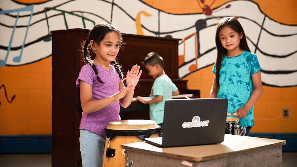 children standing in front of a laptop, one behind them playing on a piano. Wall behind them with a painted mural of floating music notation.