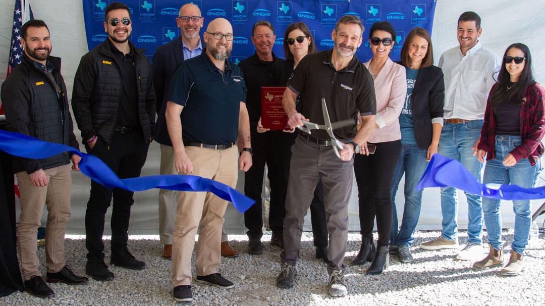 A group of people pose as one uses a large scissors to cut a blue ribbon.