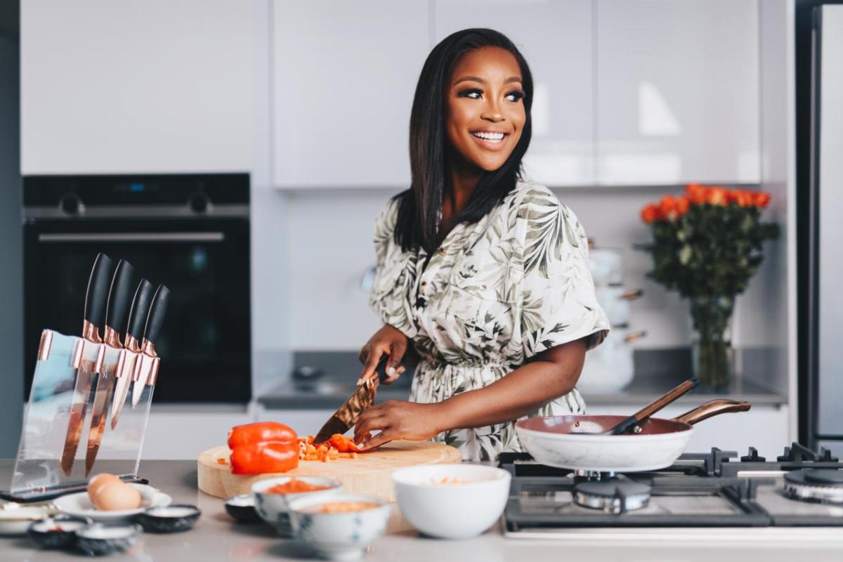 A photo of Celebrity Chef Lorna Maseko standing at a kitchen counter, looking over her left shoulder while cutting a vegetable using her new culinary line for HSN. Lorna has long black hair, medium brown skin and is smiling. She is wearing a cream and green accented short-sleeved dress.
