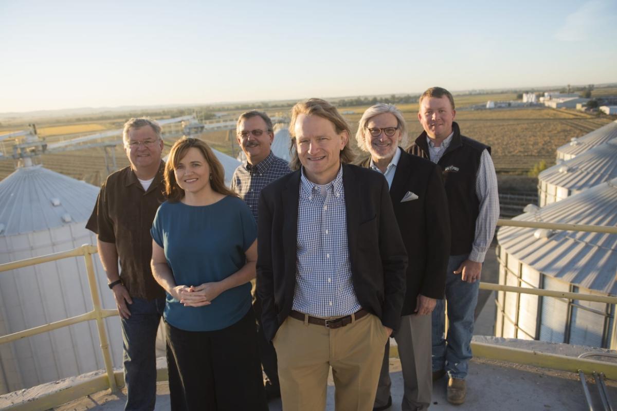 Third- and fourth-generation family members Bryce, Jessica, Mark, Grant, Tim, and Anders stand atop Lundberg’s Drying & storage facilities. Group of six people stand on a rooftop overlooking storage silos and a field