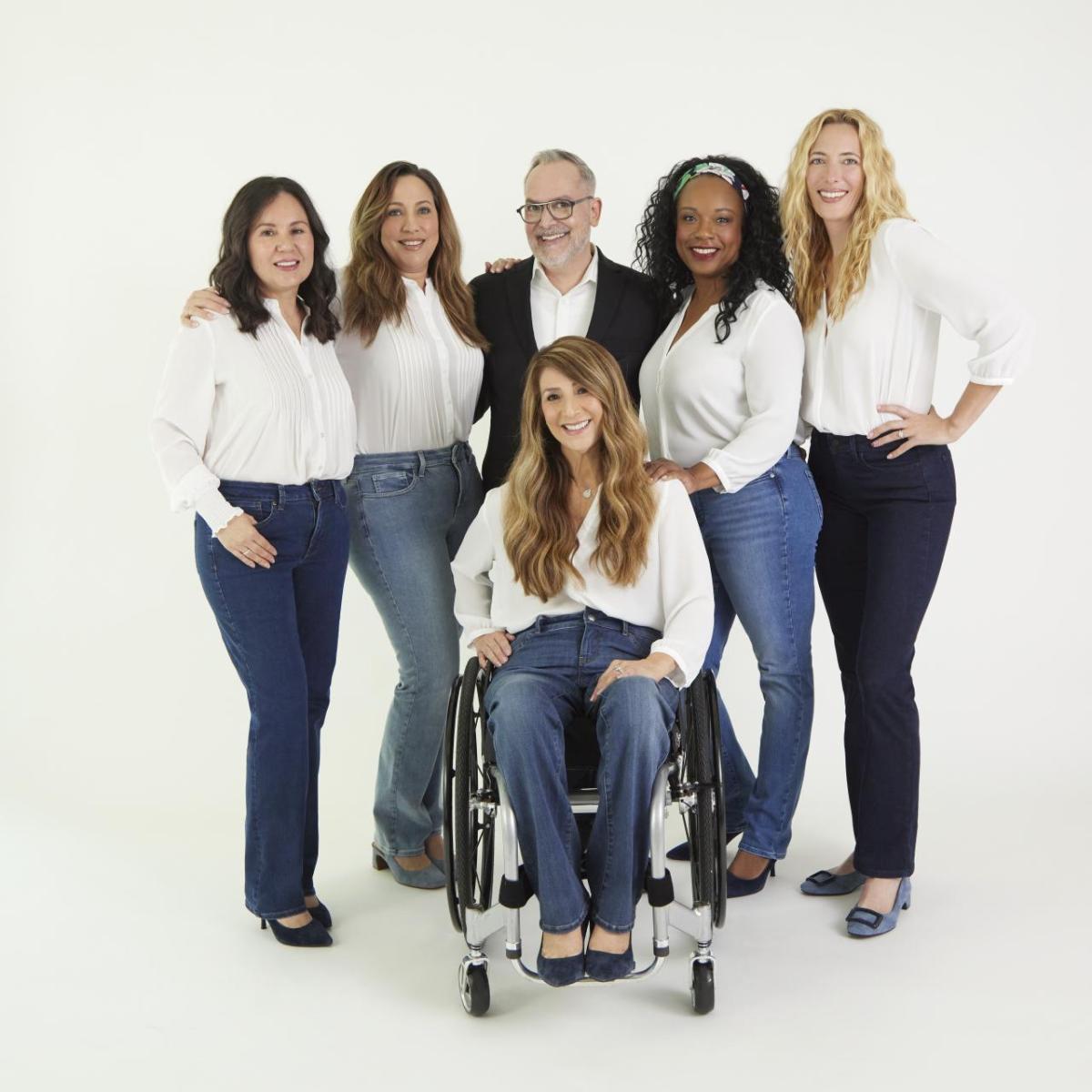 Five diverse models are showcasing NYDJ's Marilyn Straight Adapt-Denim™ jeans alongside Brand Ambassador Mark Peters. All models pose in various denim hues with white tops, while Mark, donning a white shirt and black sportscoat, stands among them. In the forefront, a wheelchair model poses elegantly.