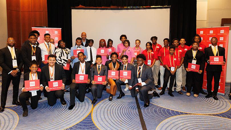 Student teams competed in the Junior Investment Club Competition during the 38th Annual Conference of the 100 Black Men of America, Inc. in Atlanta, GA. The Wells Fargo Foundation is providing a $600,000 grant to expand the Junior Investment Club to 30 markets and support other economic empowerment efforts across the U.S. Group of young people standing together at a conference holding red Wells Fargo envelopes.