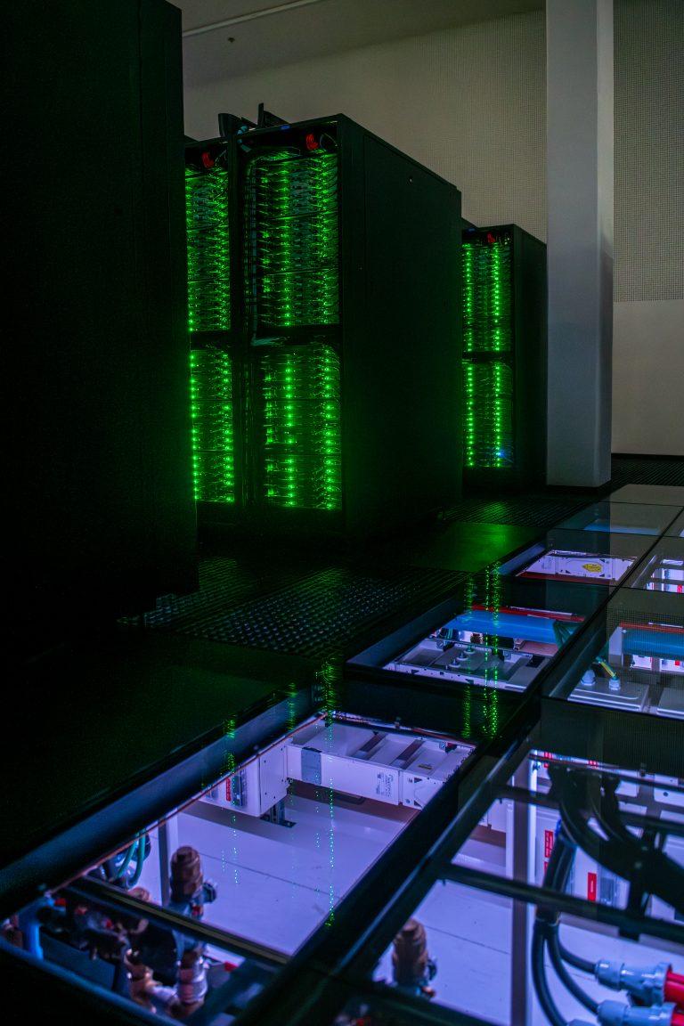 A dark upper room with green-lit large electronics. A clear section of floor showing a room below.