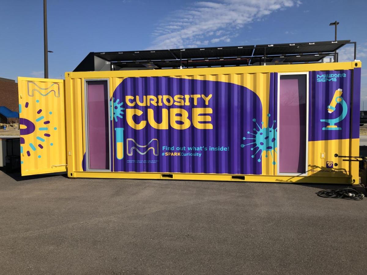 A yellow and purple shipping container with the words "Curiosity Cube" on the side showcase an environment dedicated to expanding science education