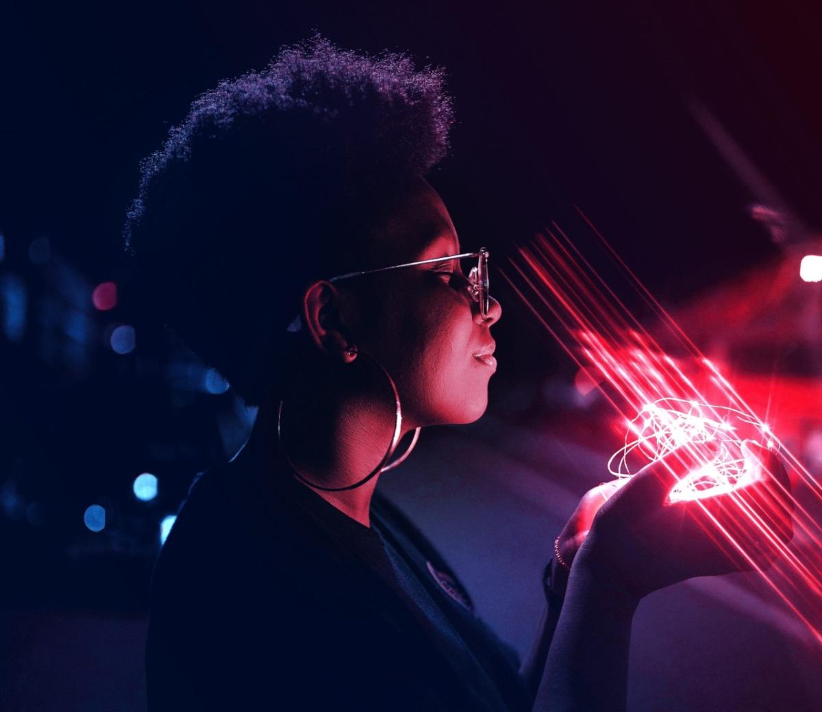 Black person with glasses and large earrings smiling with eyes closed, holding glowing pink lights