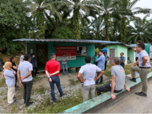 A group of people standing outside a small building surrounded by tropical trees. All listening to a speaker.