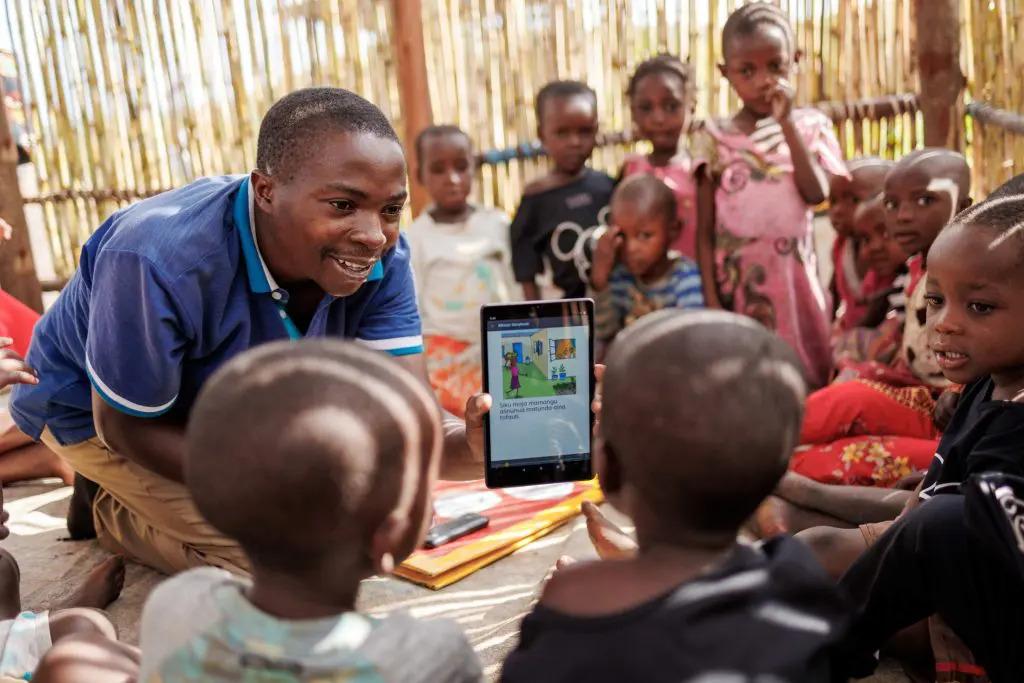 A group of children in a grass-walled hut, an adult showing them an electronic tablet with a drawing on it.