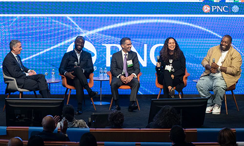 Five people seated on a stage with a huge digital display behind showing the PNC logo with blue background. All are smiling or laughing.