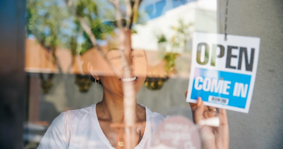 Woman in a glass window putting up an Open, Come in sign.