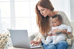 person holds a baby on their lap while working at a laptop in a home setting