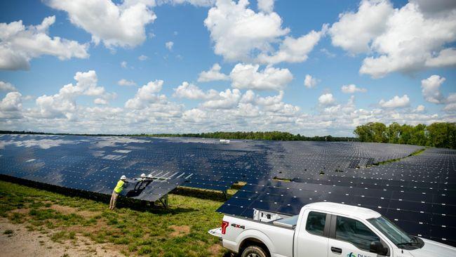 A worker inspecting a solar panel in a large field of panels. An open cloudy sky above.