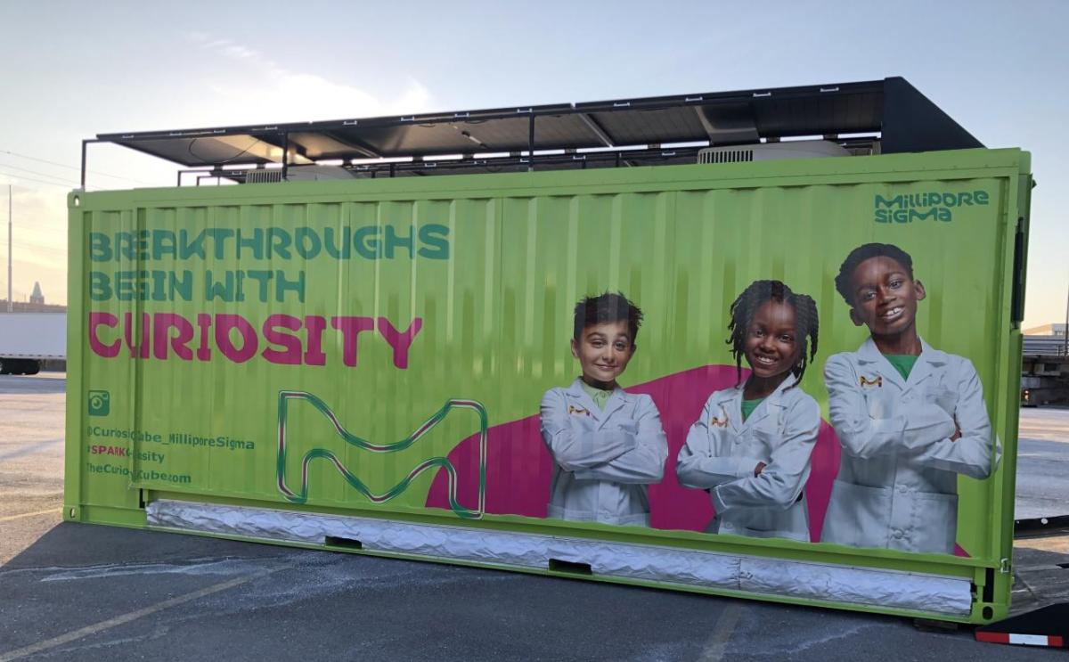 A green shipping container turned mobile science lab features three young scientists and text reading, "Breakthroughs Begin With Curiosity."