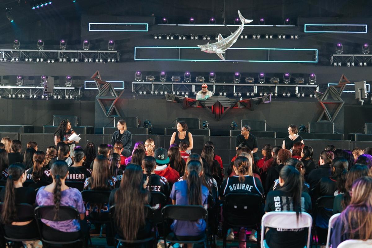 A panel of speakers sits on stage facing a large seated audience inside a festival-style venue, with stage lighting rigs and a sculptural element hanging above.