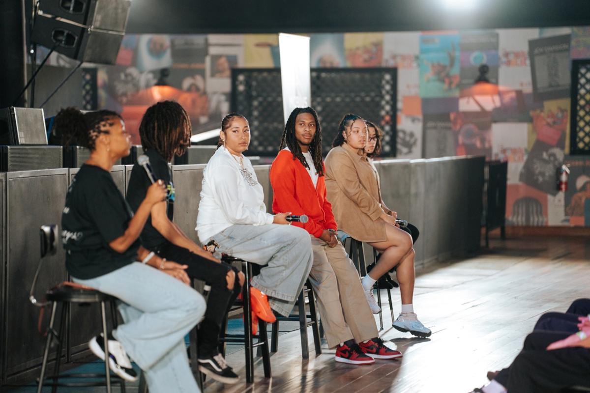 Six panelists sit on stools on a stage holding microphones, facing an audience off-camera, with venue artwork and lighting visible in the background.