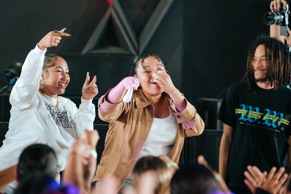 Three performers stand at the front of a stage engaging with a crowd; one holds a microphone while others gesture toward the audience, with hands raised in the foreground.