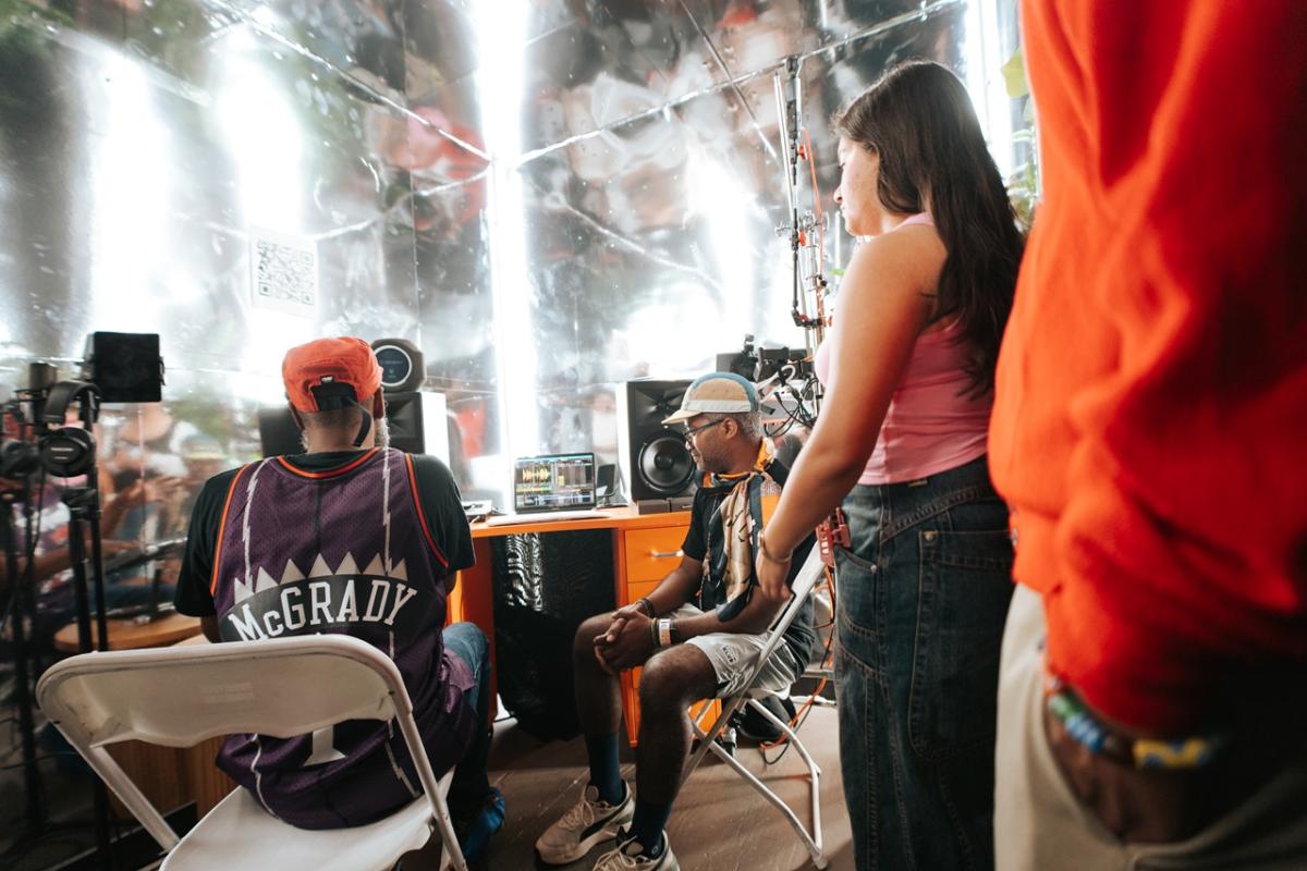Several people sit and stand around audio and video equipment inside a reflective booth, looking toward a laptop screen displaying sound or video controls.