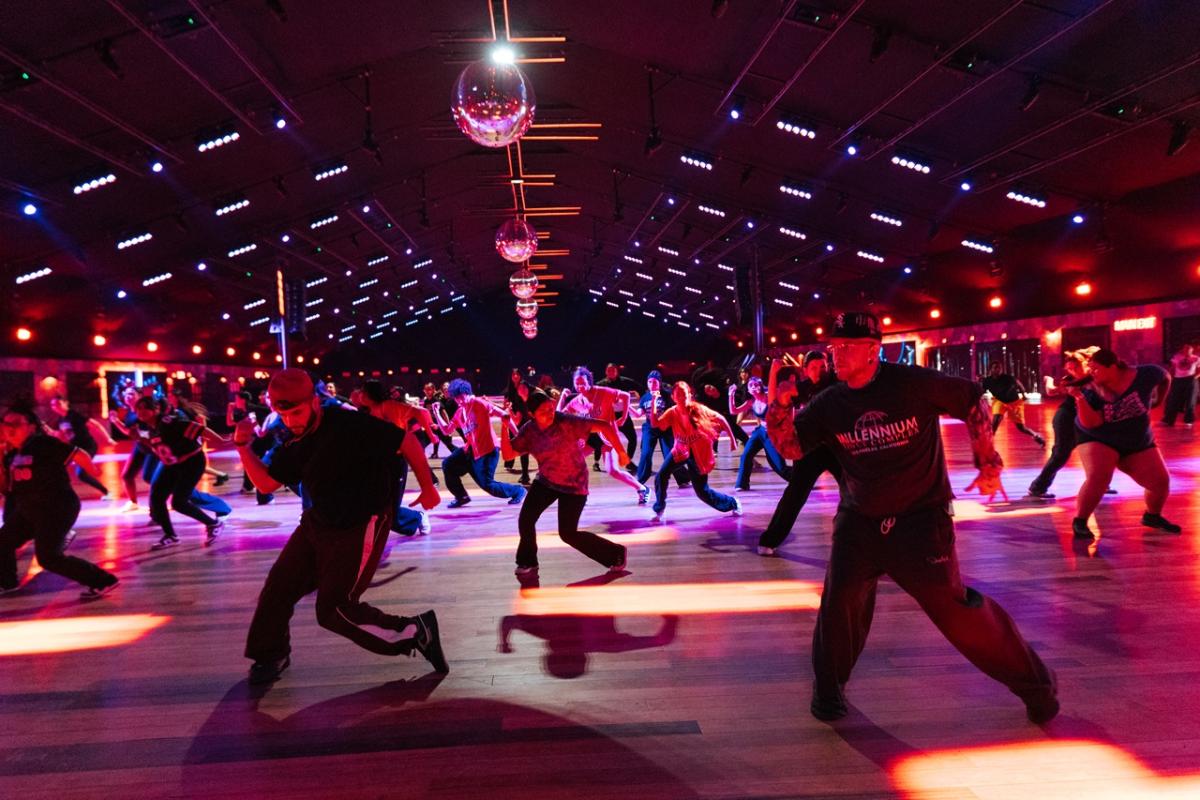 A large group of dancers moves across a wide dance floor under colorful lights and disco balls inside an indoor venue, captured mid-routine.