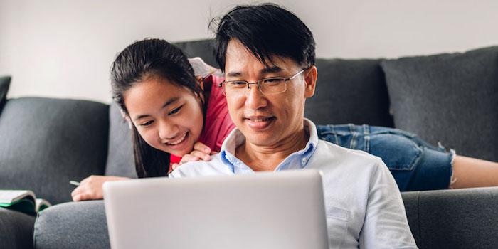 Father and daughter (daughter is sitting on a couch and dad is sitting on the floor) are looking at a laptop screen 