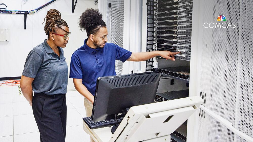 Two people in a room full of computer servers, a screen and keyboard on a small table in front of them.