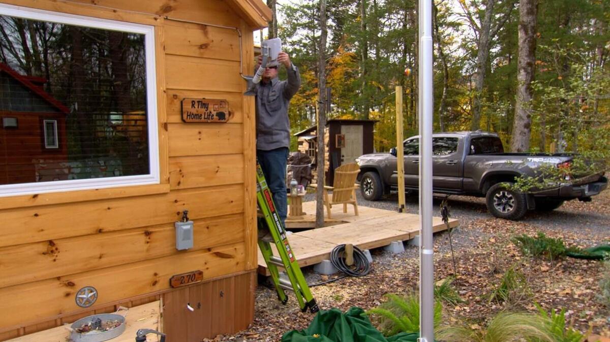 a person installing equipment on the side of a home in a wooded area. A grey truck behind them.