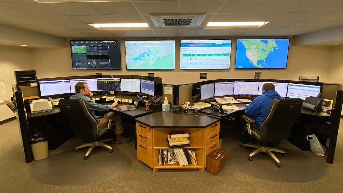 two people seated at desks with multiple monitors in front of them and mounted on the wall in front of them. Maps and data showing on the screens.